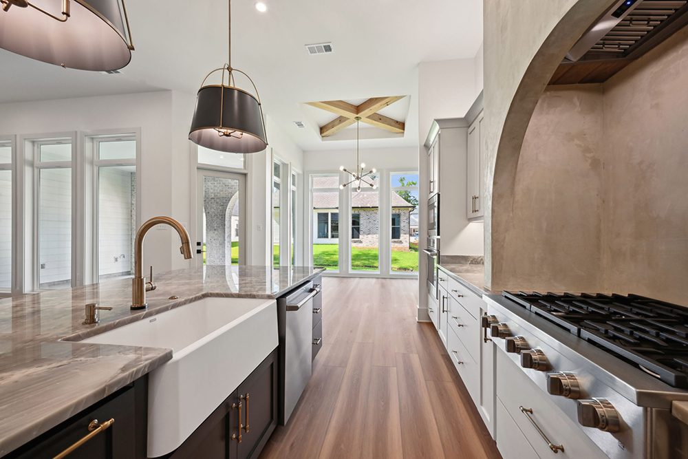 This kitchen captures the perfect blend of luxury craftsmanship and open-air beauty. A large central island with a stunning gray stone countertop features a deep farmhouse sink, sleek brushed gold faucet, and built-in dishwasher—ideal for prepping meals while enjoying the surrounding elegance. Overhead, modern black-and-gold pendant lights add a refined statement. To the right, a professional-grade gas cooktop is nestled beneath a dramatic arched plaster hood, creating an architectural focal point that’s both rustic and refined. The custom white cabinetry paired with polished hardware completes the gourmet aesthetic, while double ovens and ample counter space add functionality.