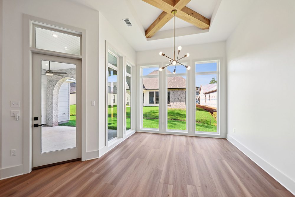 Above, a vaulted tray ceiling is highlighted by exposed natural wood beams and a contemporary sputnik-style chandelier, adding character and a modern statement piece. Wide-plank wood flooring enhances the warm, welcoming ambiance and ties in beautifully with the neutral palette of the home. Whether used for everyday meals or elegant gatherings, this dining area provides a bright, open space with style, sophistication, and a connection to the outdoors.