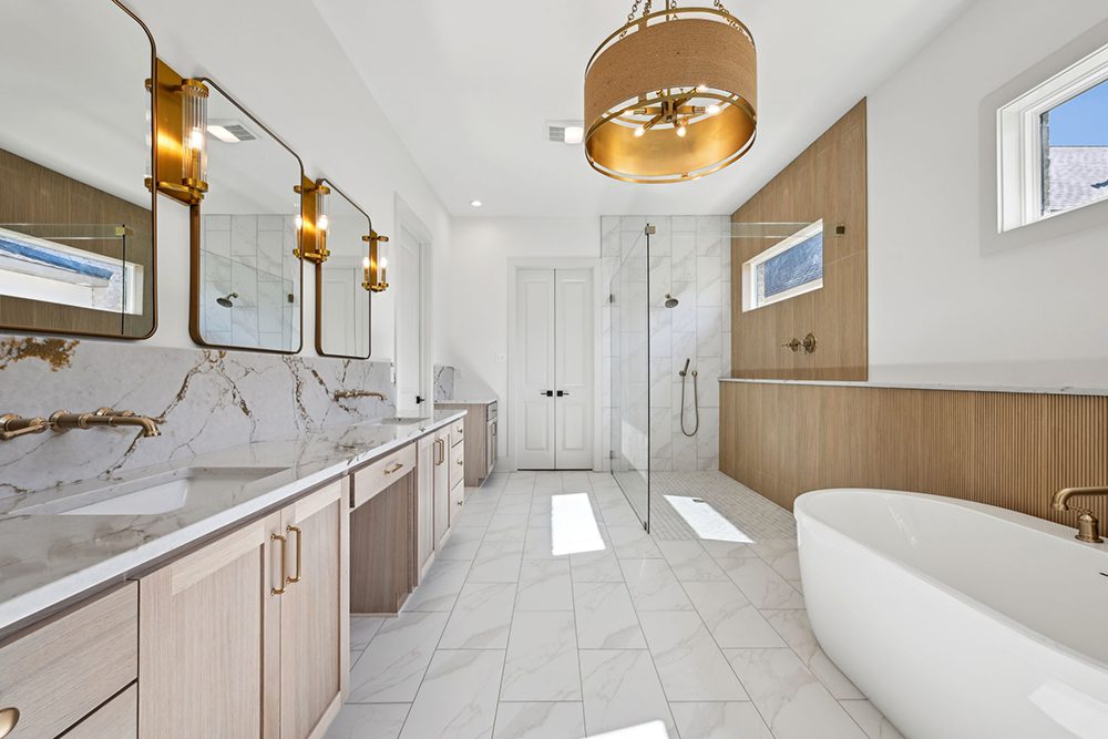 Overhead, a statement light fixture with a fabric and gold drum design enhances the spa-like ambiance. Bright natural light pours in through multiple windows, highlighting the clean lines and serene color palette of white, gold, and light wood. Every detail in this bathroom is thoughtfully curated to create a calming and luxurious retreat for everyday indulgence.