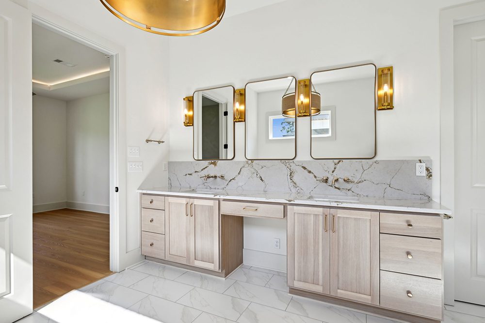 This secondary vanity space perfectly complements the primary bathroom with a design that balances luxury and practicality. Framed by three bronze-trimmed mirrors and illuminated by sleek vertical sconces, the vanity provides an ideal setup for personal care routines. The central section offers a seated makeup station, while both sides feature ample storage with soft wood cabinetry and brushed gold hardware.