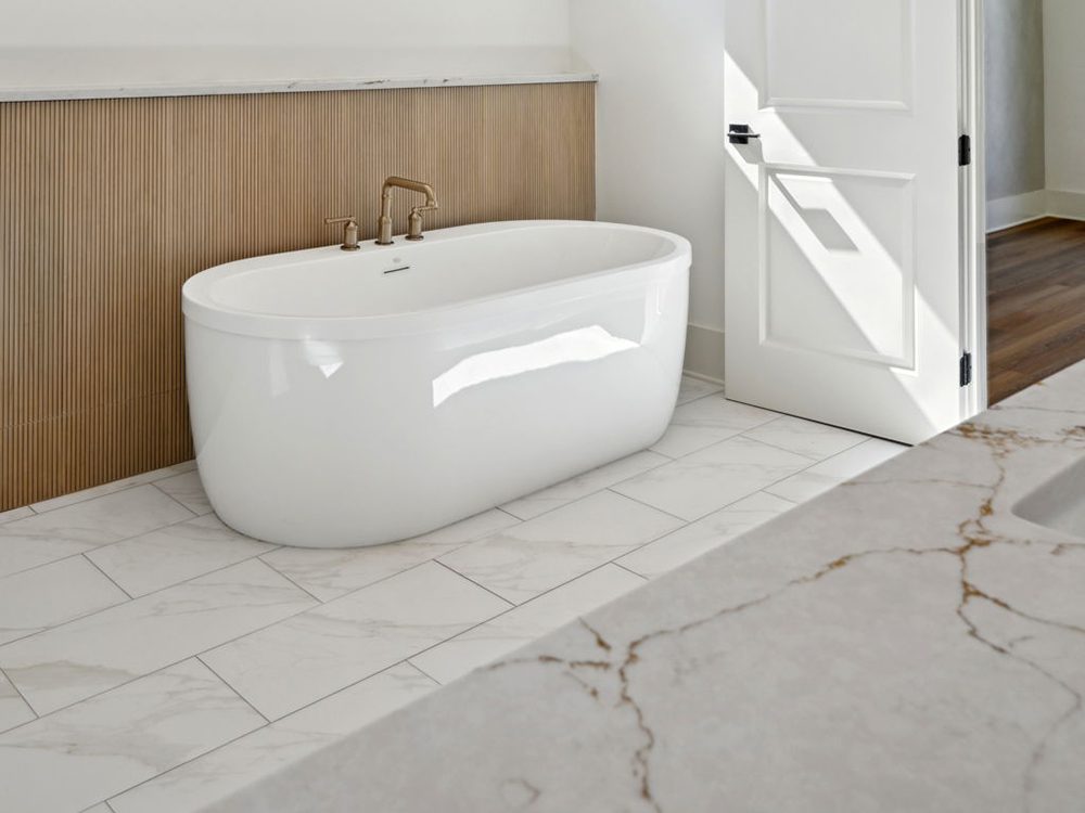 This serene bathroom moment captures a freestanding soaking tub that exudes modern simplicity and comfort. The tub’s clean, curved silhouette sits against a textured wood-paneled wall detail, adding warmth and organic contrast to the cool white finishes.