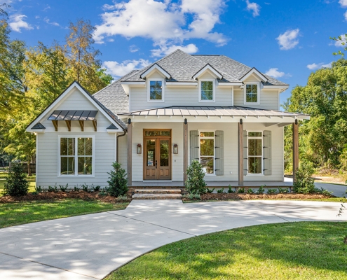 Windows are adorned with decorative shutters and trim, and the left side has a charming gabled section with exposed wood brackets under a smaller metal awning. The house is surrounded by a manicured lawn and neat landscaping, with shrubs lining the foundation, while the curved driveway leads up to the home under a clear, blue sky.