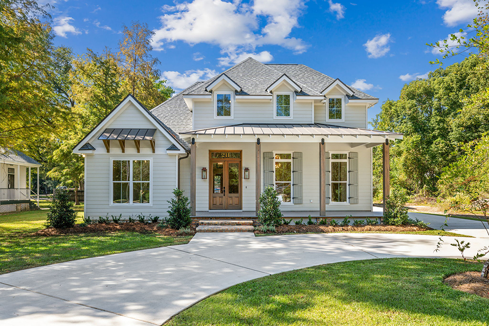 Windows are adorned with decorative shutters and trim, and the left side has a charming gabled section with exposed wood brackets under a smaller metal awning. The house is surrounded by a manicured lawn and neat landscaping, with shrubs lining the foundation, while the curved driveway leads up to the home under a clear, blue sky.