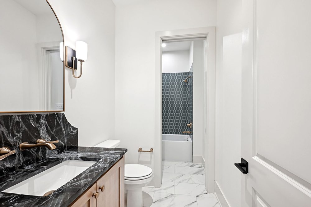 This chic guest bathroom features a bold, black marble vanity with dramatic white veining, paired with matte gold fixtures and a soft wood-toned cabinet base. A curved-edge mirror and dual sconces offer a balanced glow, complementing the sleek design.