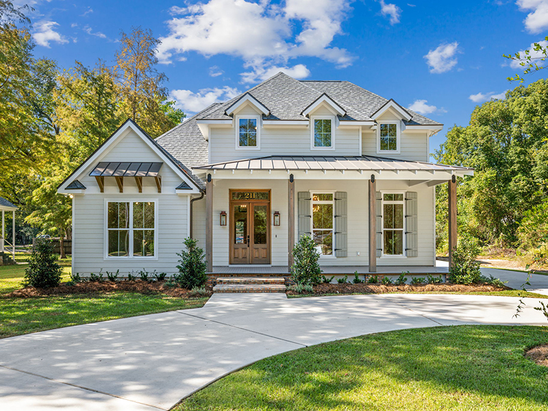 The image shows a charming two-story house with a classic design, featuring light-colored siding and a gray shingle roof. The front elevation has a welcoming covered porch supported by wooden columns, with a metal awning and brick steps leading up to a double wooden front door flanked by glass panels.