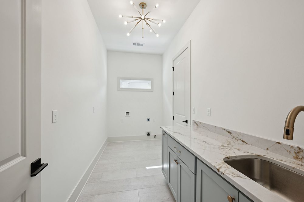 This bright and spacious laundry room combines functionality with a modern aesthetic. A long quartz countertop with elegant gold veining provides ample workspace, complemented by muted sage cabinetry and a sleek undermount sink with a brushed gold faucet.