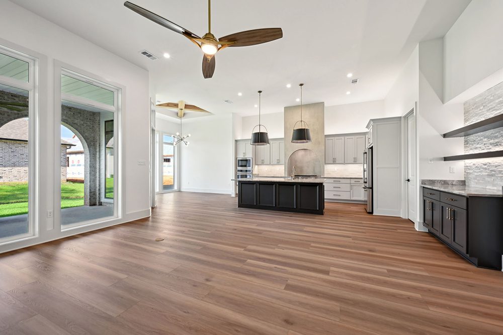 This open-concept kitchen and living area boasts warm wood flooring, oversized windows offering abundant natural light, and a dramatic modern ceiling fan.