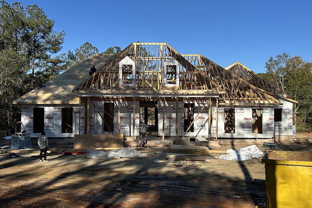This photo captures a home under construction, showcasing its framework and roof structure in progress. The exterior walls are wrapped in Tyvek house wrap for weatherproofing, while exposed wooden trusses outline the roof and dormer windows.